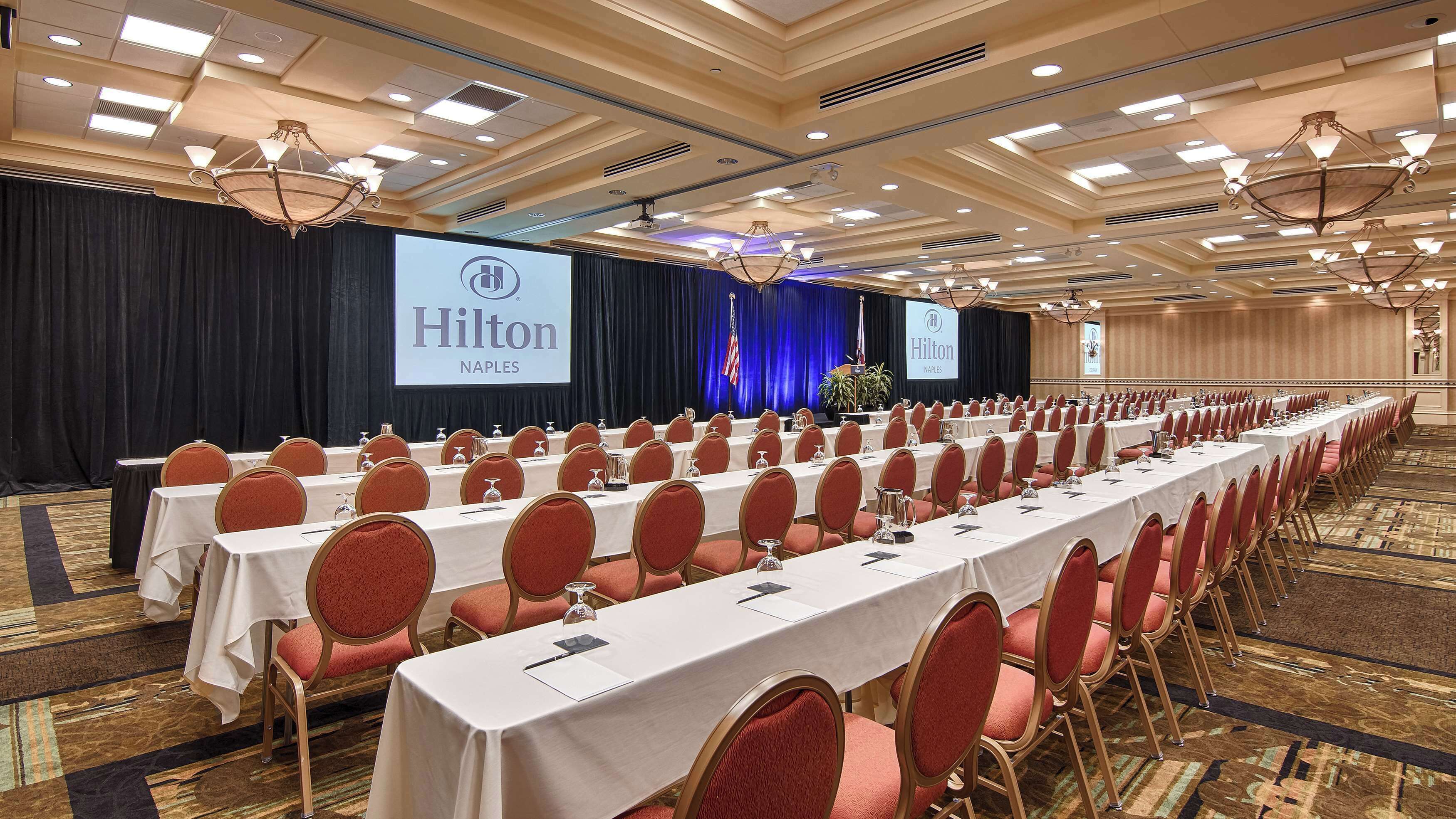 A conference room at the Hilton Naples hotel with rows of tables and chairs, a stage with a large screen, and a chandelier.