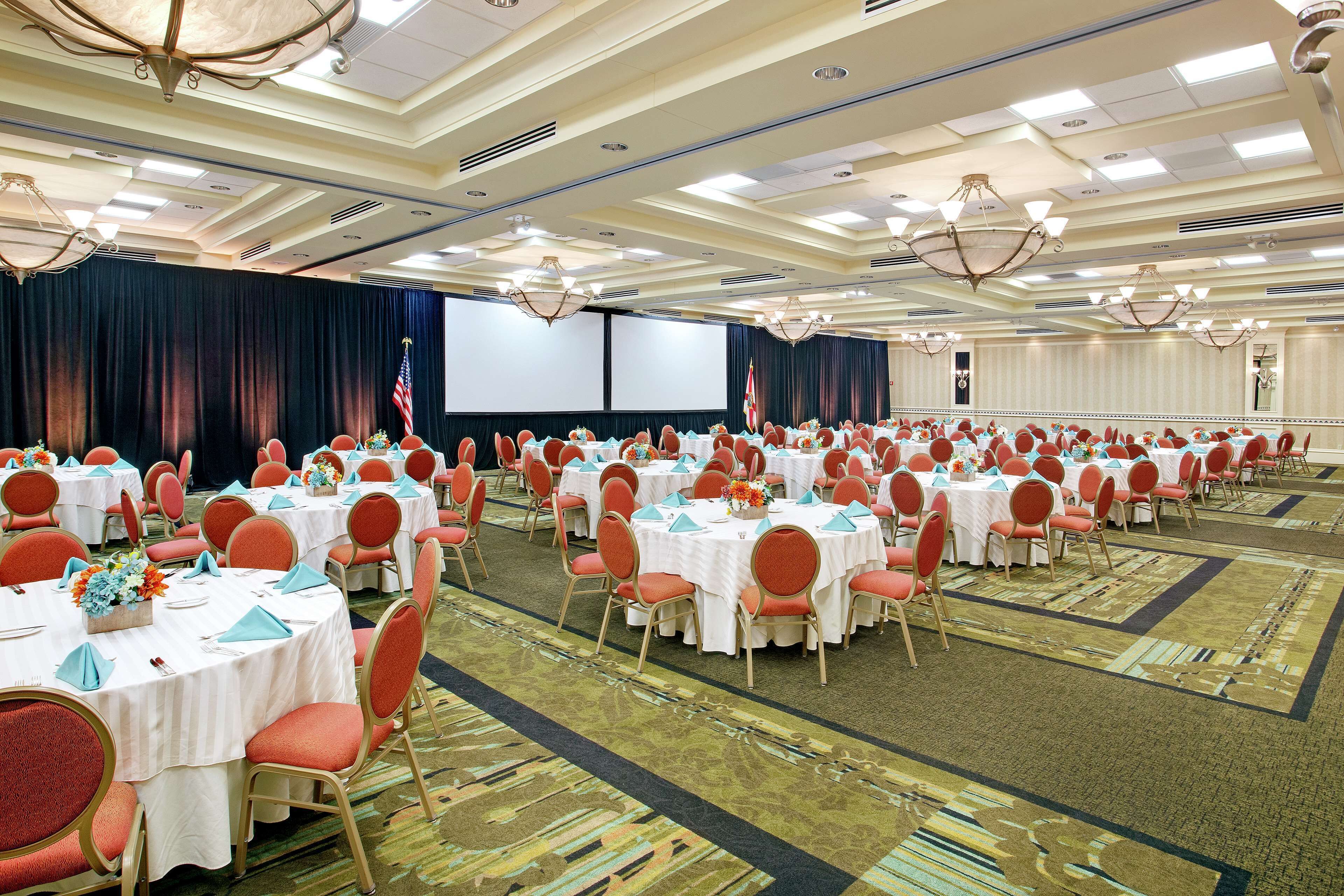 A large conference room with rows of tables and chairs, a stage with a screen, and an American flag.