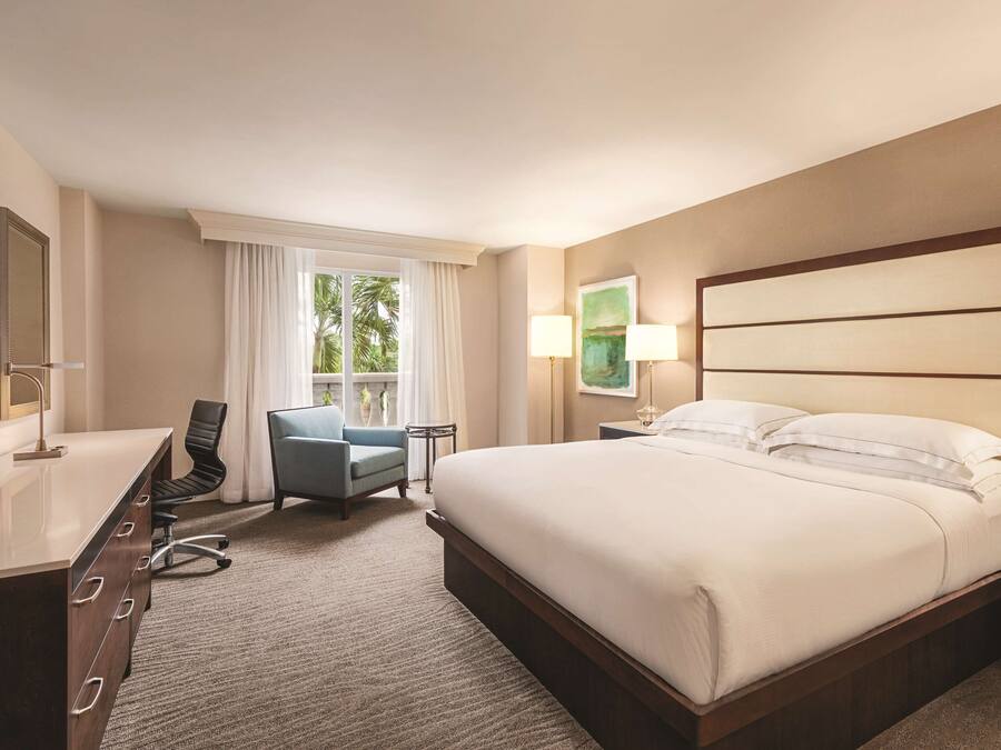 Ein Hotelzimmer mit einem großen Bett, einem Schreibtisch, einem Stuhl und einem Fenster mit Vorhängen.