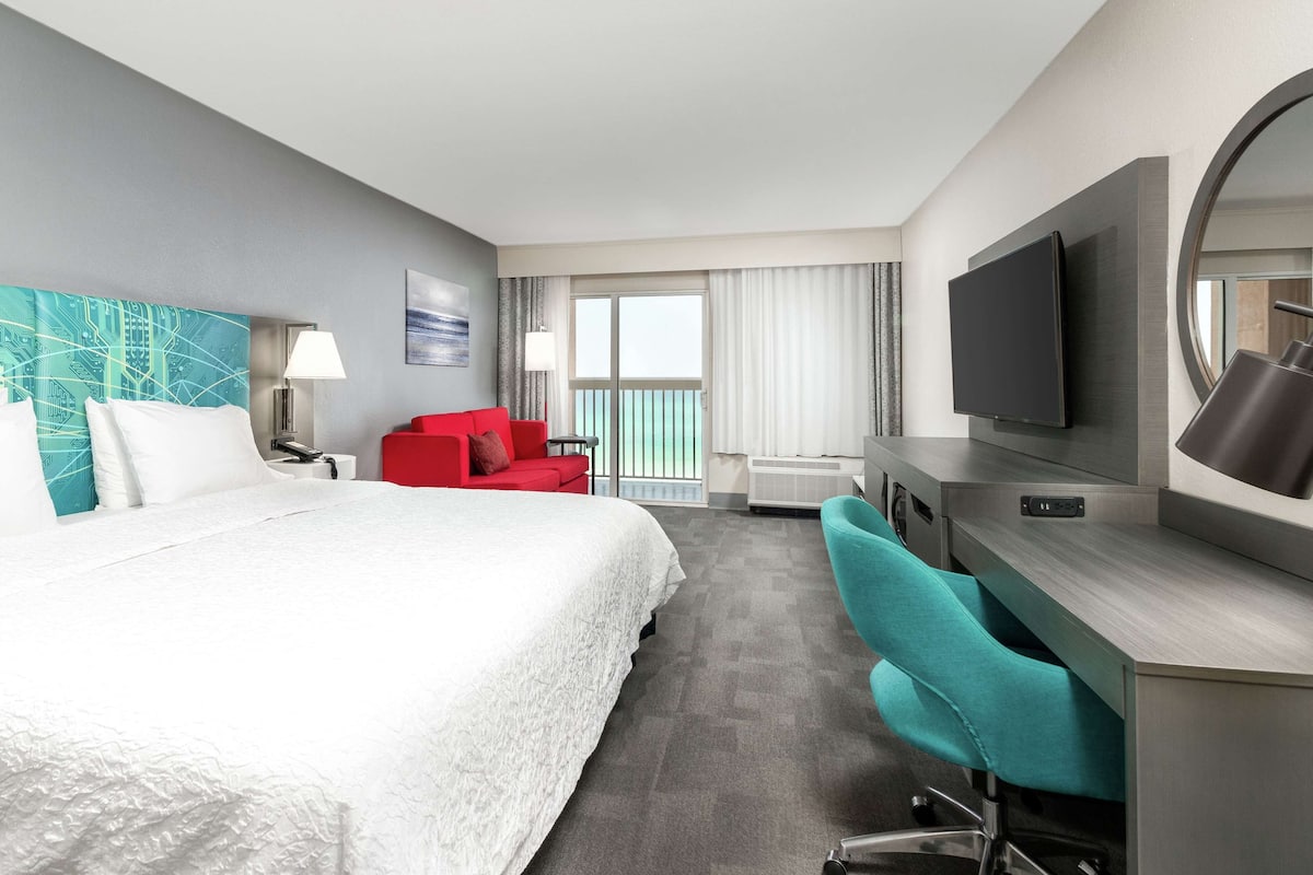 A hotel room with a large bed, a desk with a TV, a red chair, and a window with curtains.