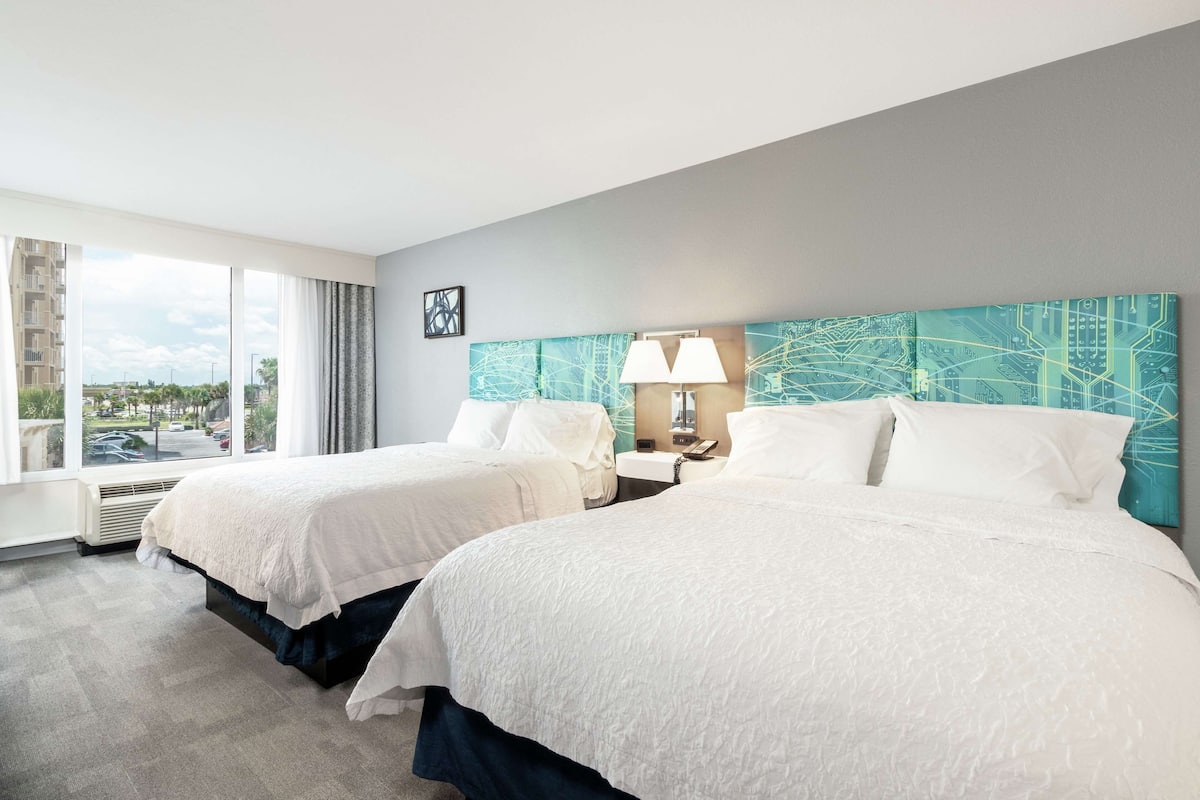 A hotel room with two beds, a large window with a view, a modern headboard, and a bedside table with a lamp.