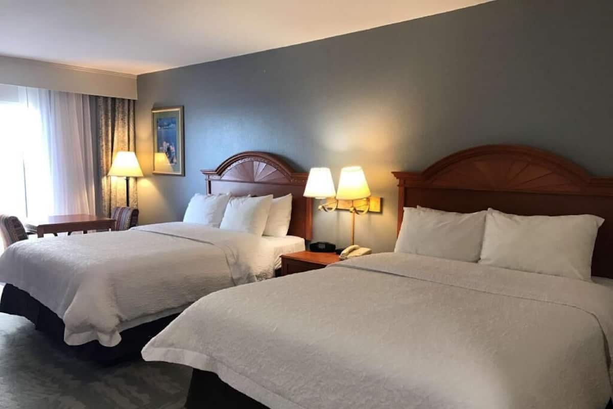 A hotel room with two beds, a desk, a chair, and a lamp.