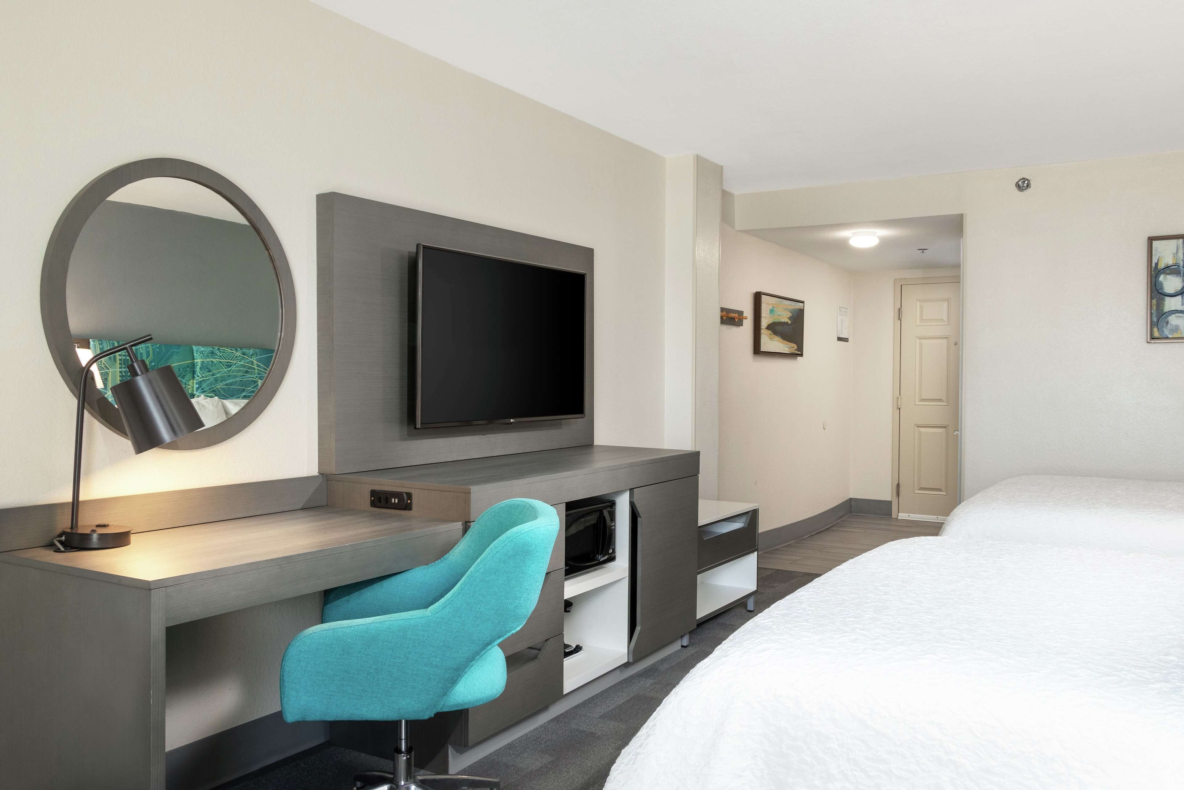 A hotel room with a bed, a desk with a chair, a television, a mirror, and a door.