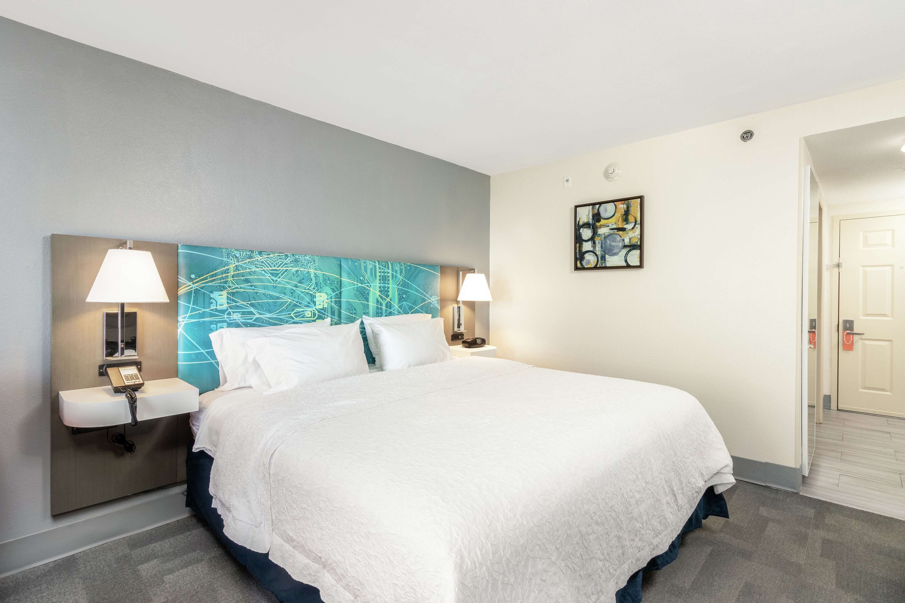 A hotel room with a large bed, two bedside lamps, a nightstand with a phone, a blue accent wall, and a painting on the wall.