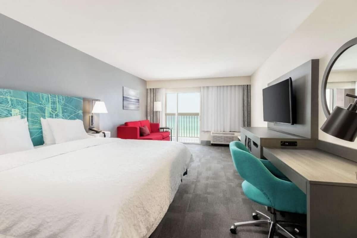 A hotel room with a bed, a desk with a chair, a TV, a red chair, and a large window with curtains.