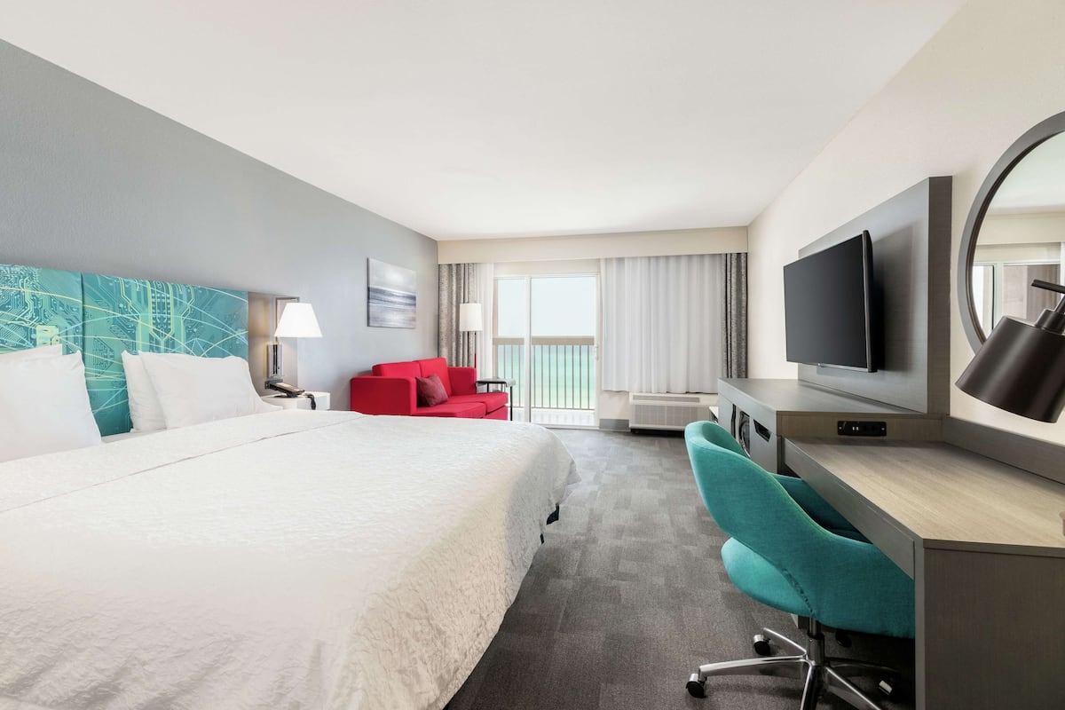 A hotel room with a large bed, a desk with a TV, a red chair, and a view of the outdoors.