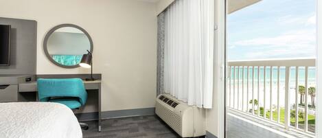 A hotel room with a bed, a desk with a chair, a round mirror, a heater, and a balcony with a view of the beach.