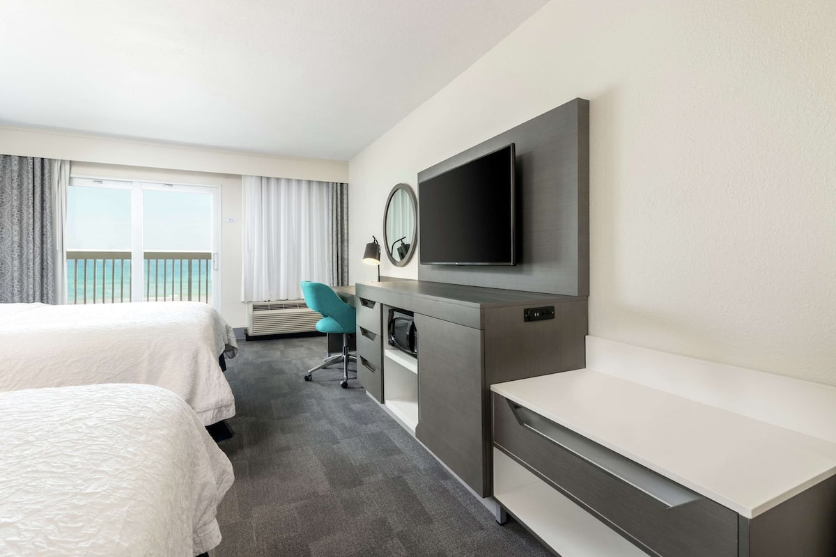 A hotel room with two beds, a desk, a chair, a flat-screen TV, and a balcony with curtains.