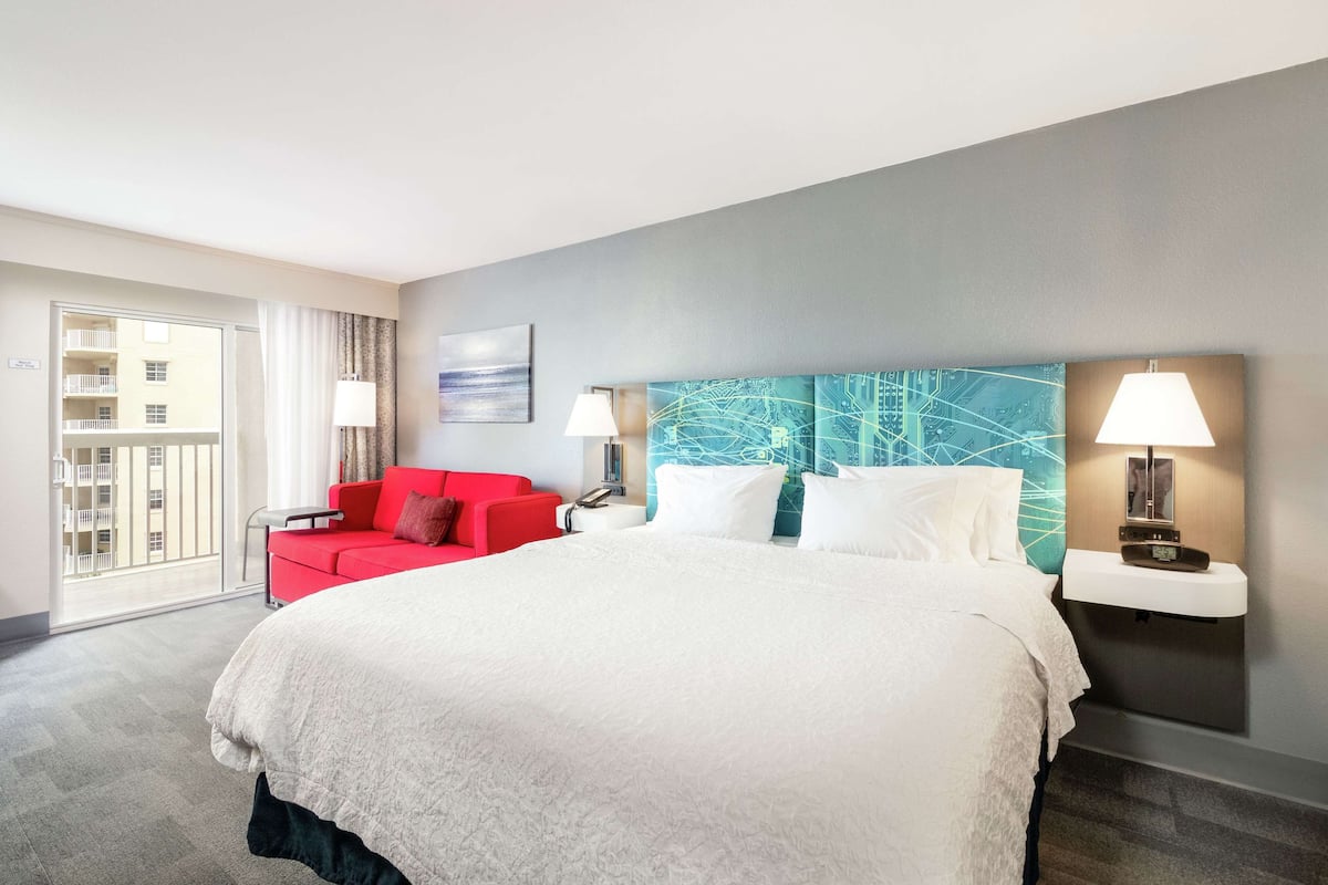 A hotel room with a large bed, a red sofa, a wall-mounted lamp, and a view of the outside balcony.