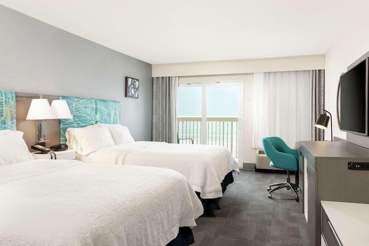 A hotel room with two beds, a desk, a TV, and a balcony with curtains.