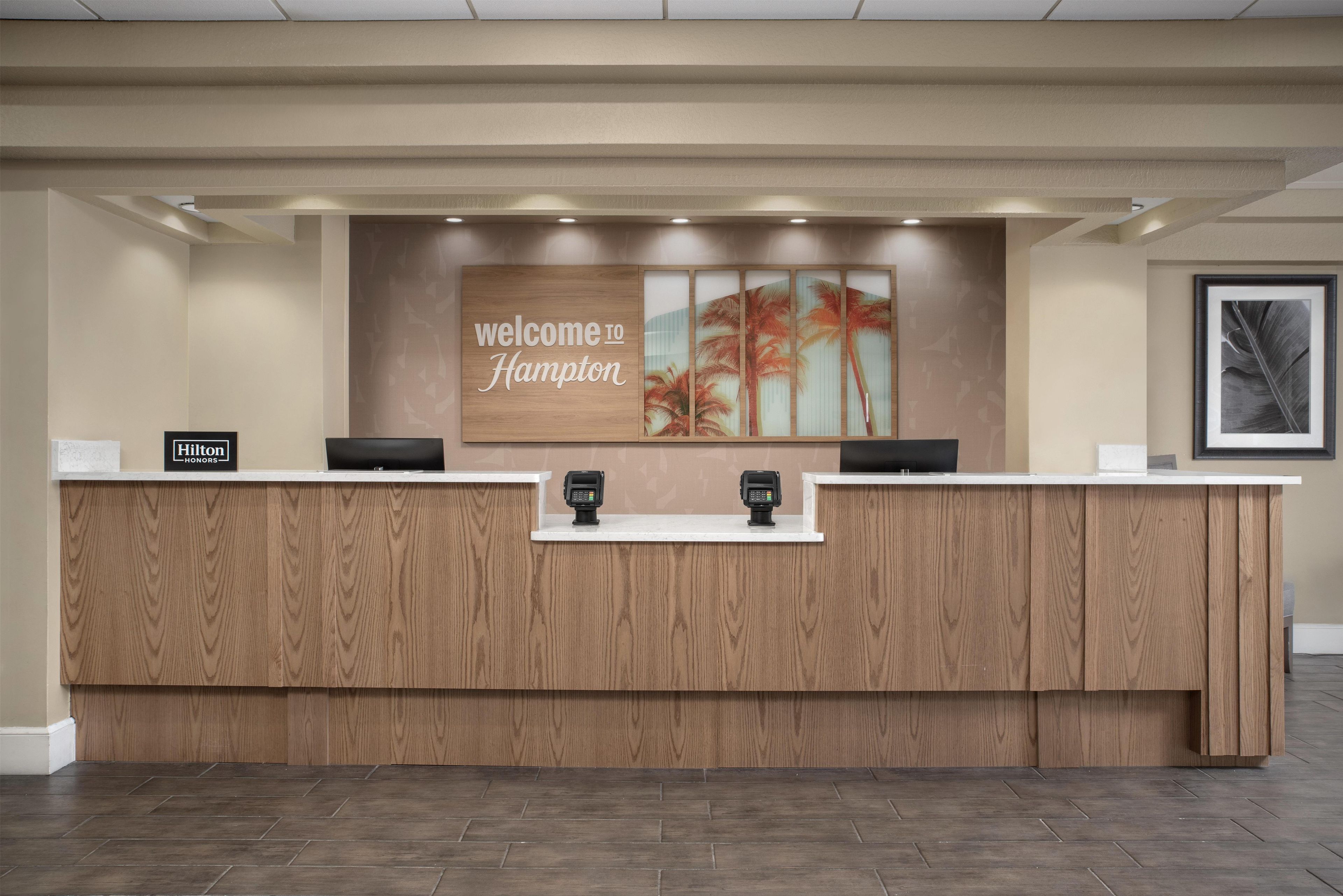 A reception desk with a 'welcome to Hampton' sign, two card readers, and a 'Hilton Honors' plaque.