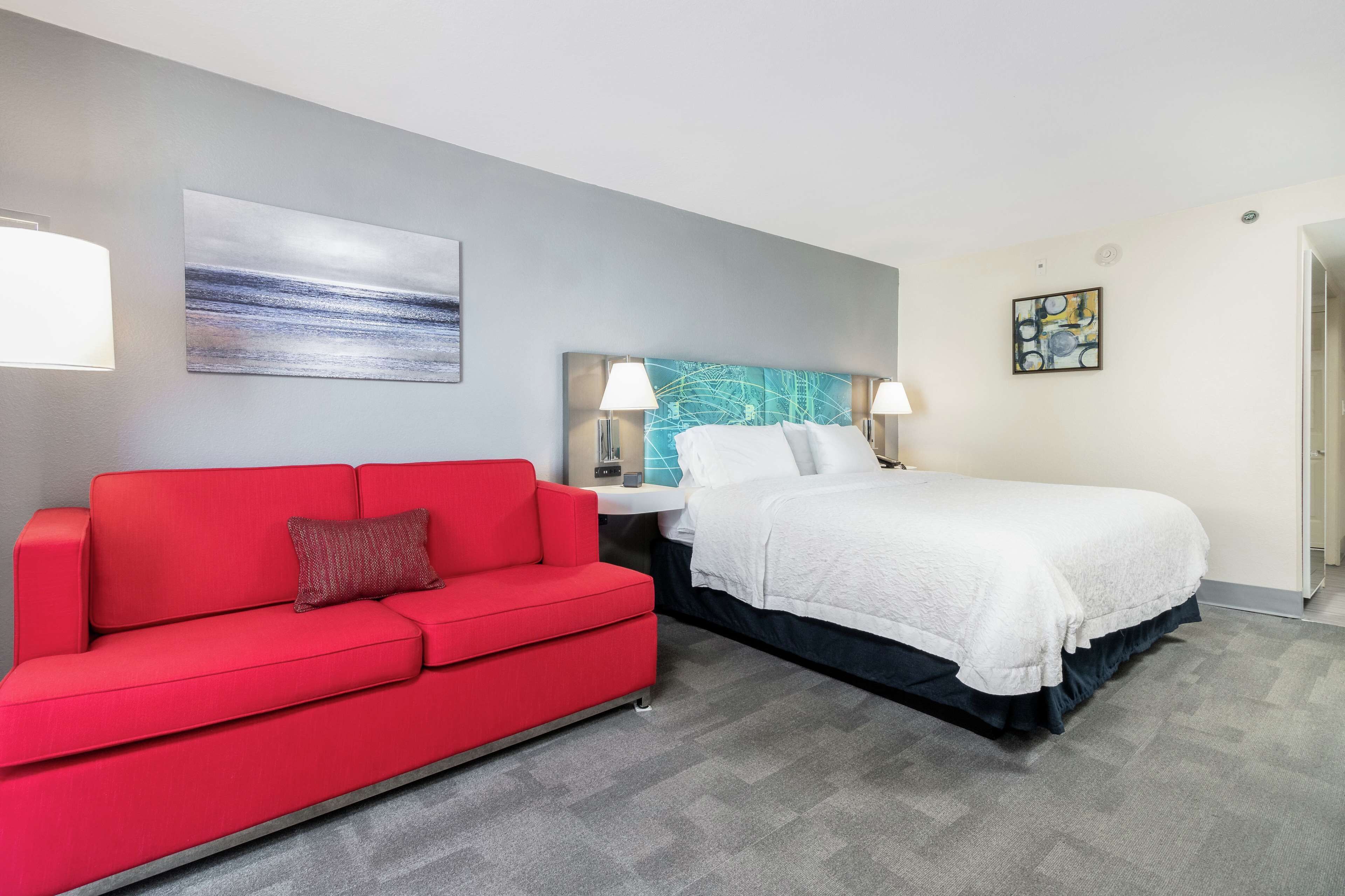 A hotel room with a red sofa, a bed with white bedding, a blue headboard, and a painting on the wall.