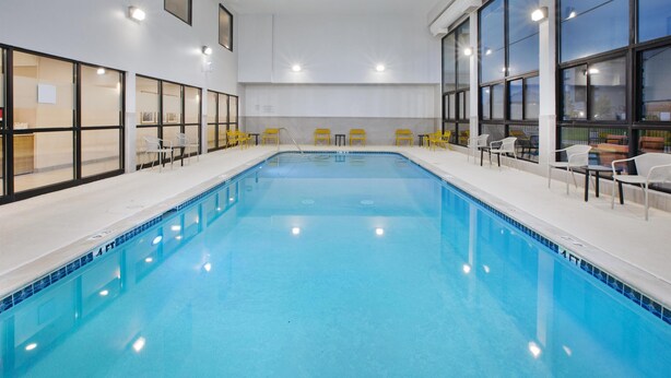 An indoor swimming pool with clear blue water, surrounded by yellow chairs and tables, large windows, and a white interior. - Hampton Inn Hotel Detroit/Belleville-Airport Area (Belleville)