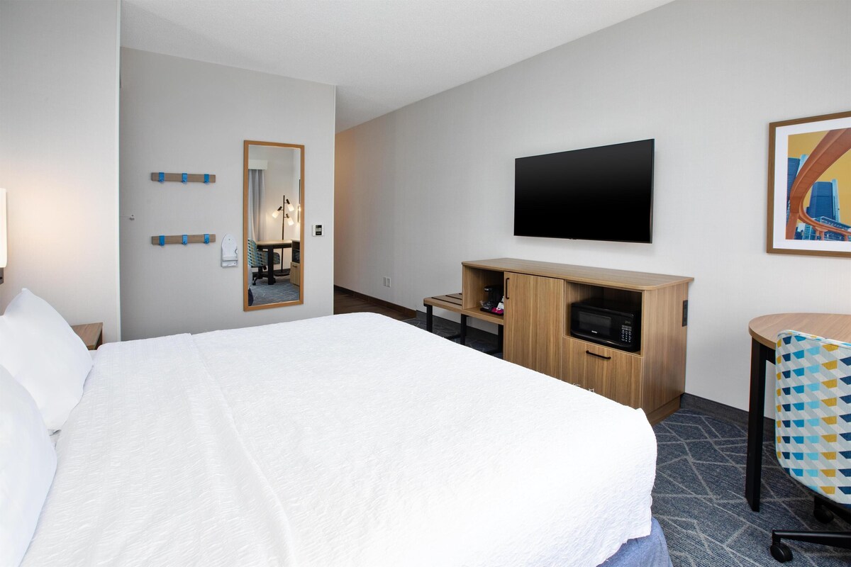 A hotel room with a large bed, a wooden desk, a flat-screen TV, a mirror, and a chair.