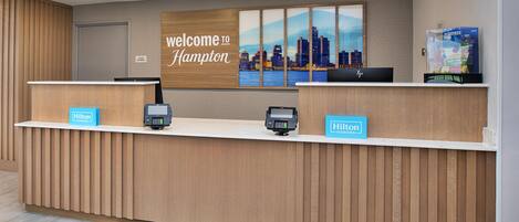 A hotel reception desk with a 'Welcome to Hampton' sign, a cityscape poster, and a 'Hilton Honors' loyalty card display.
