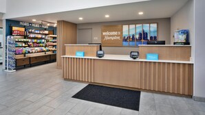A hotel reception area with a 'Welcome to Hampton' sign, a snack bar with various packaged foods, and a digital check-in station. - Hampton Inn Hotel Detroit/Belleville-Airport Area (Belleville)