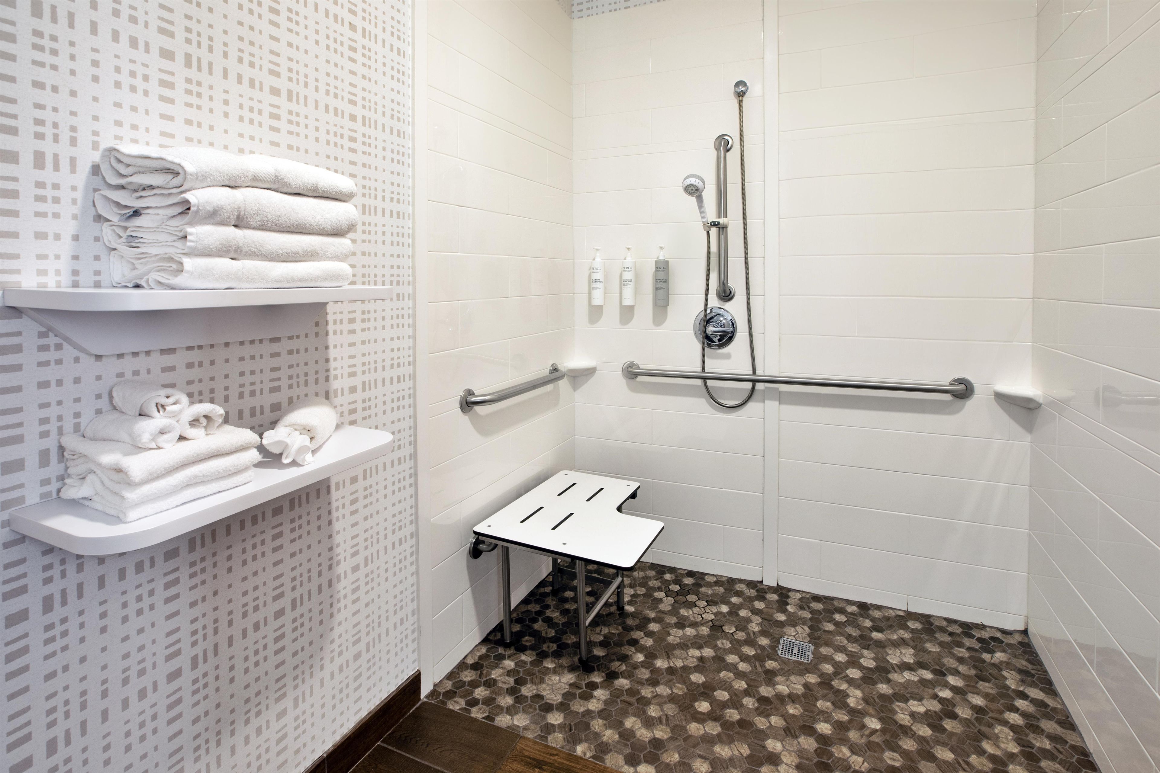 Un área de ducha accesible, con pared de azulejos blancos, estante con toallas dobladas y piso con diseño hexagonal.