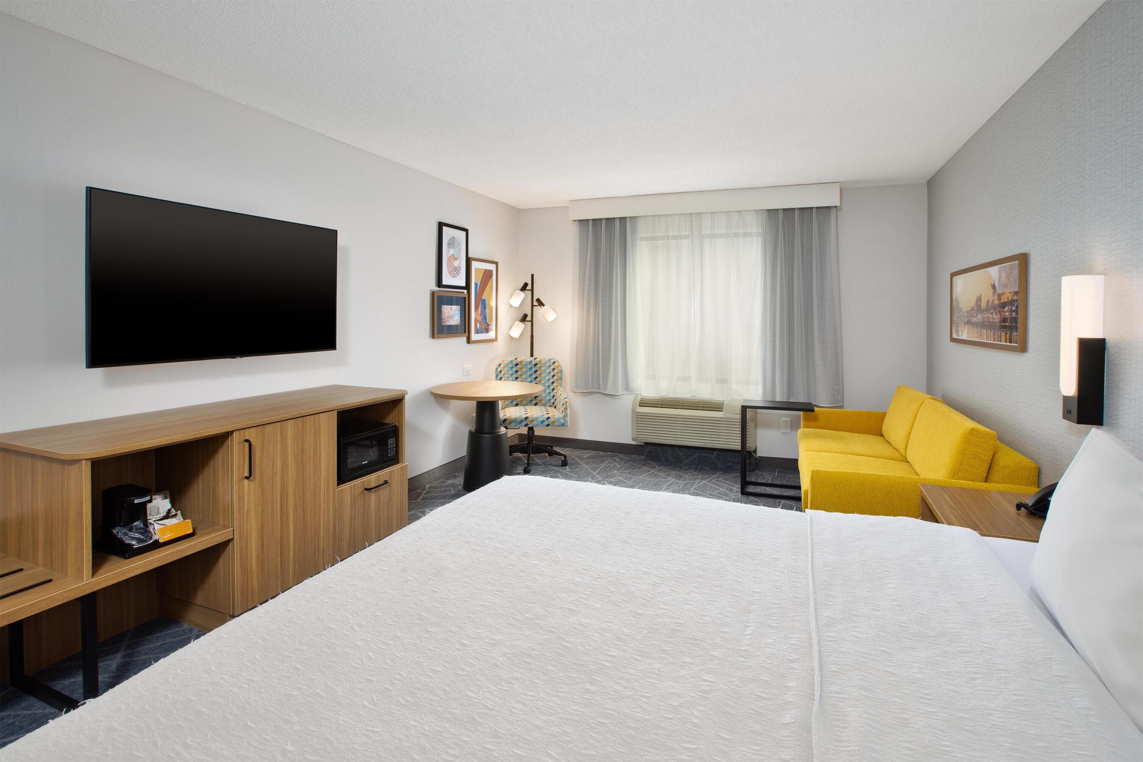 Una camera d'albergo con un letto grande, una scrivania con una sedia, un tavolino, un divano, una TV a schermo piatto e una finestra con tende.