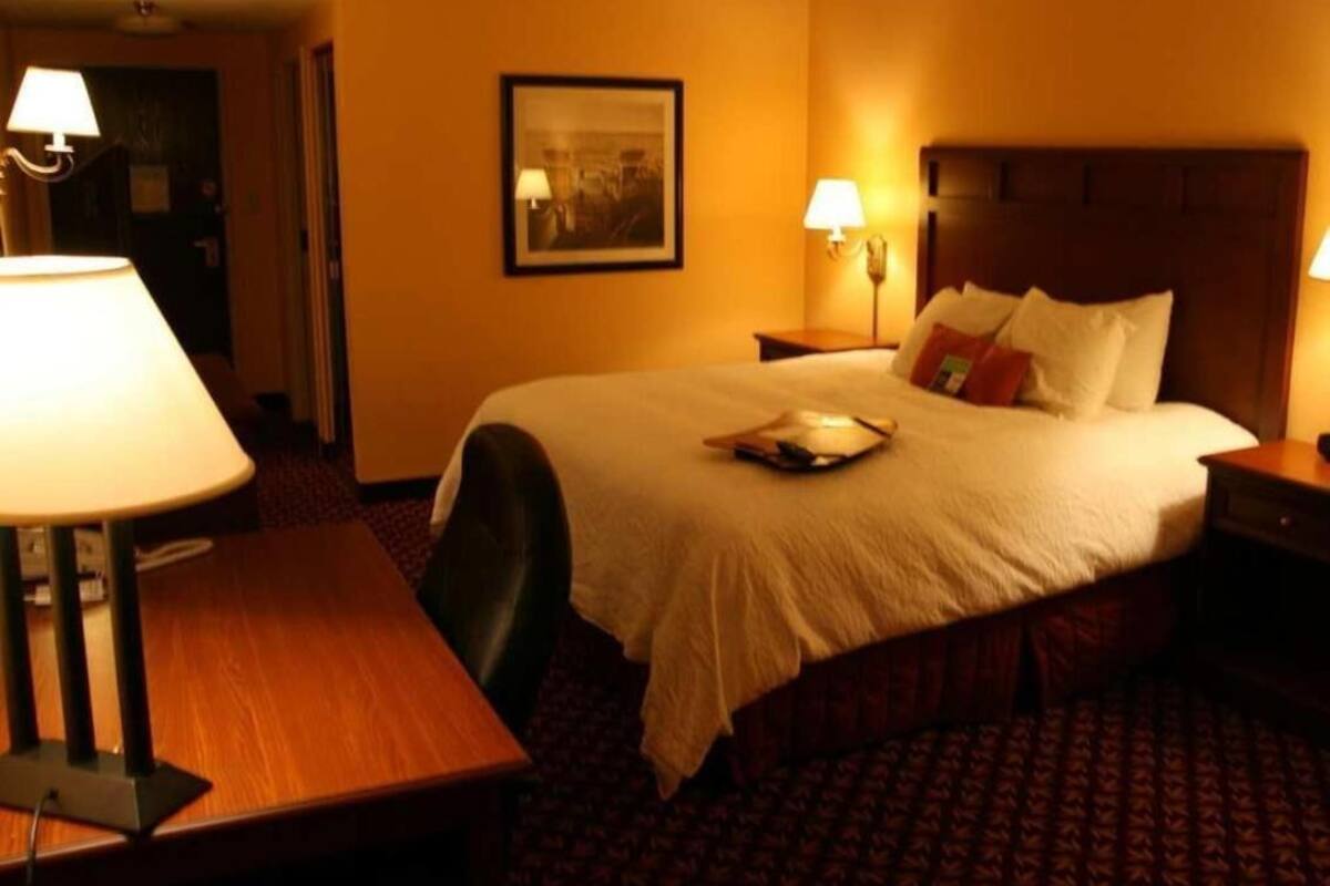 A hotel room with a bed, a desk, a chair, a lamp, a framed picture on the wall, and a nightstand.
