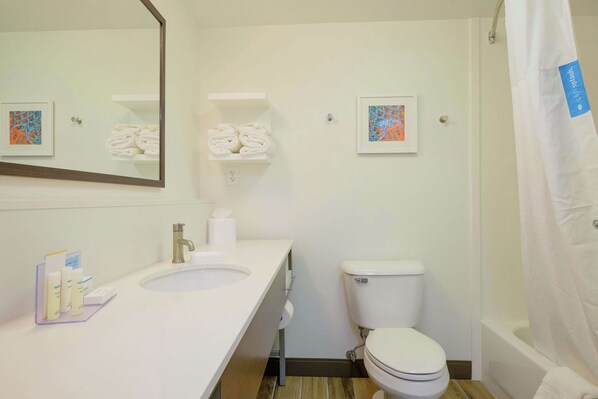 Un baño con lavamanos blanco, inodoro y cortina de ducha. Hay dos cuadros enmarcados en la pared y un estante con toallas dobladas.
