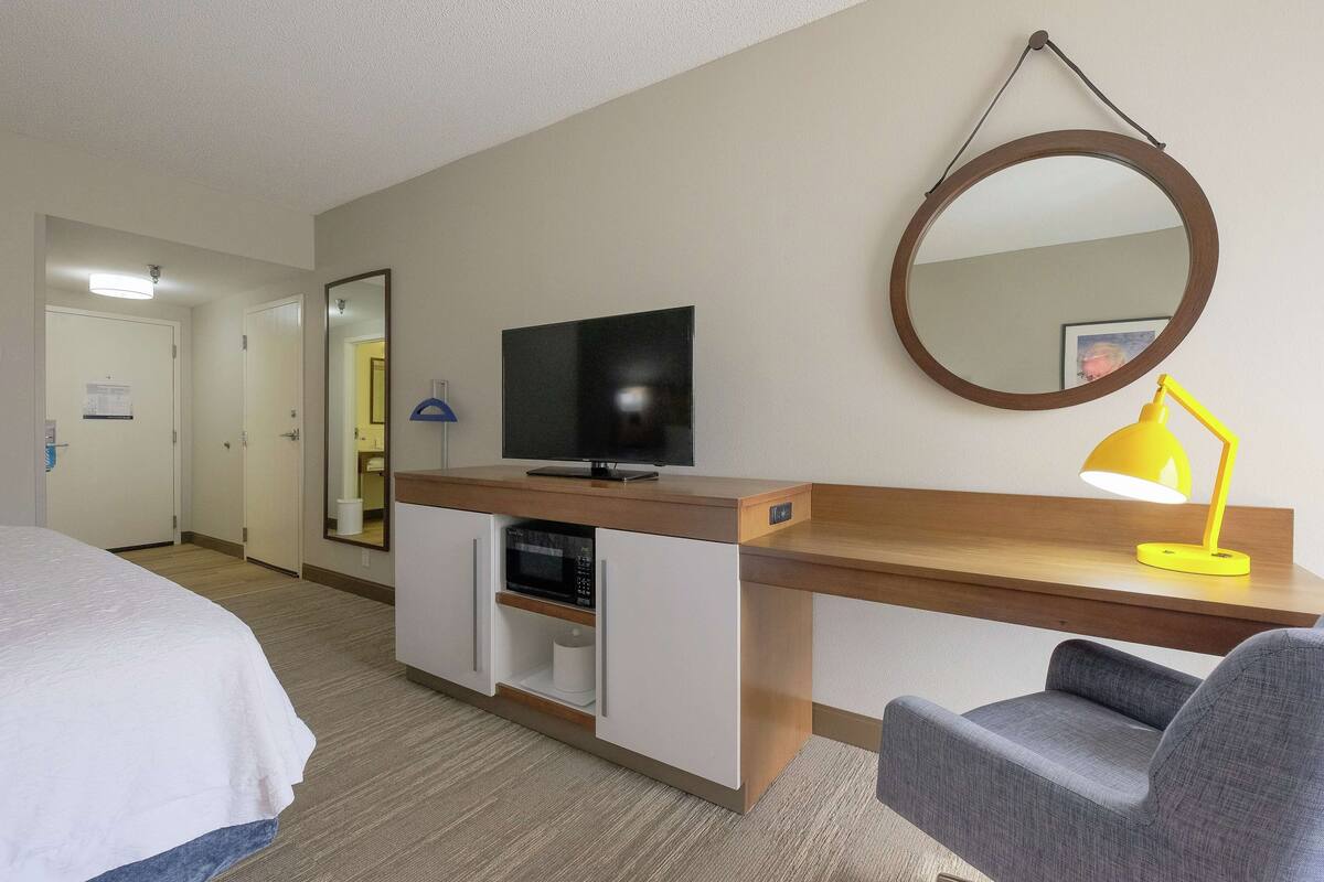 Habitación de hotel con una cama, un escritorio con televisión y lámpara, un microondas y un espejo.