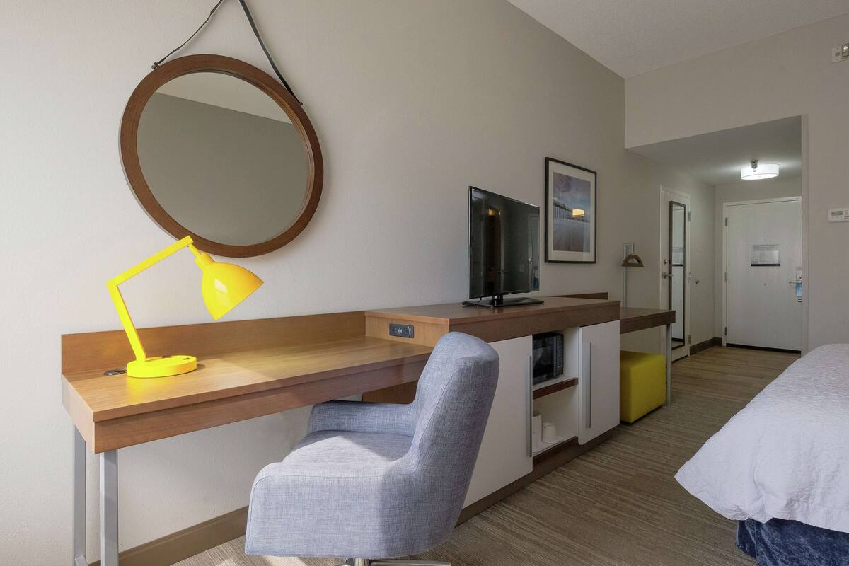Habitación de hotel con escritorio de madera, una silla, televisor, cama, espejo y puerta.