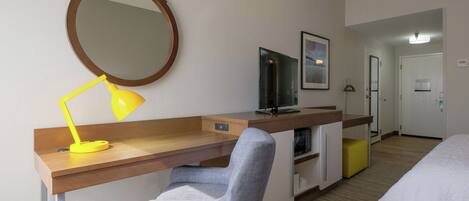 Habitación de hotel con escritorio de madera, una silla, televisor, cama, espejo y puerta.