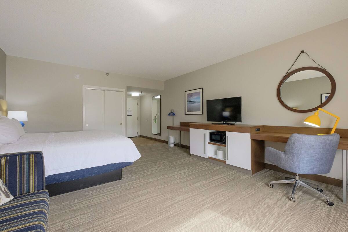 Habitación de hotel con cama, escritorio, computadora, silla, televisor y espejo.