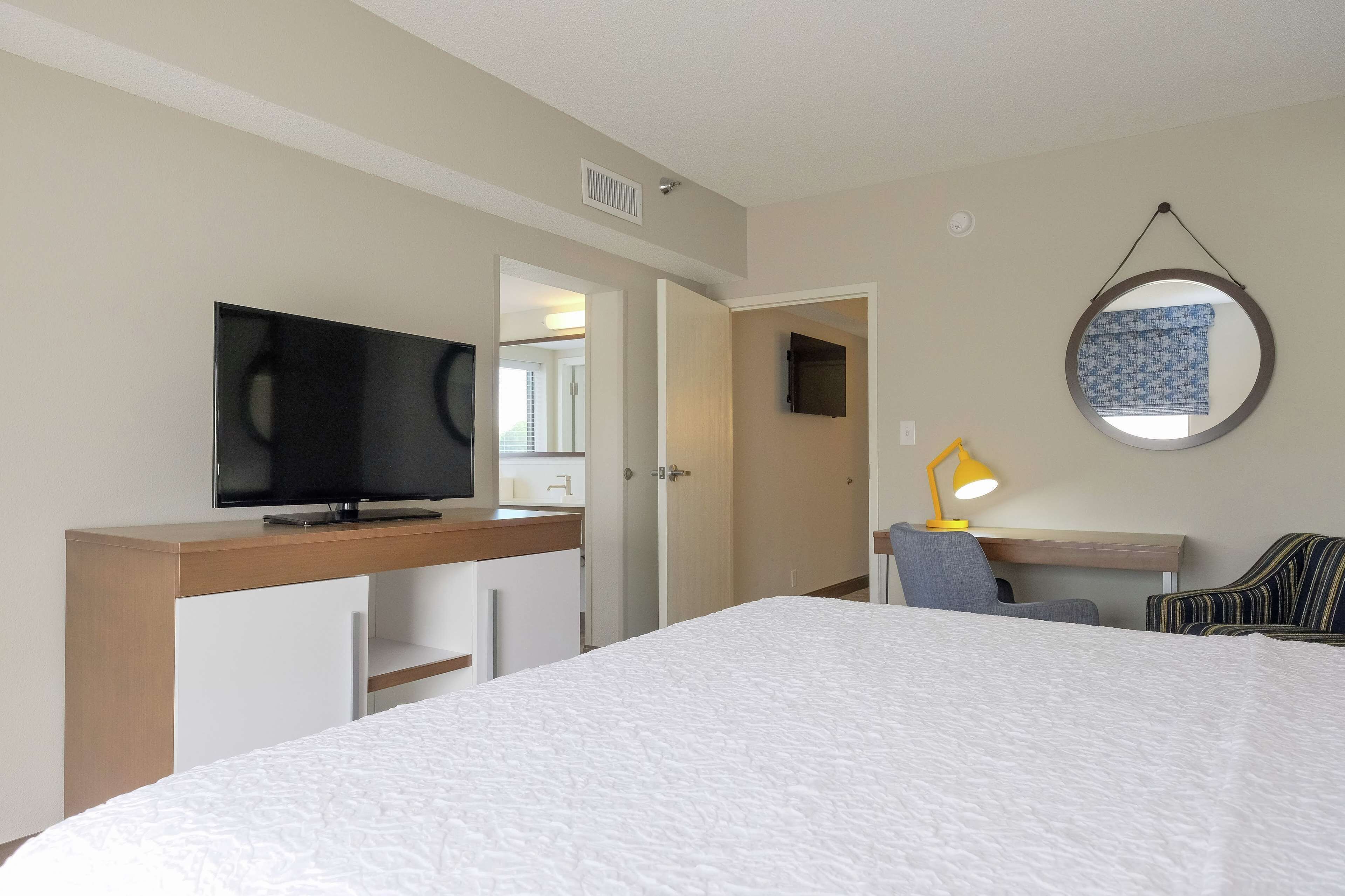 A hotel room with a bed, a desk with a television, a chair, a lamp, and a mirror on the wall.