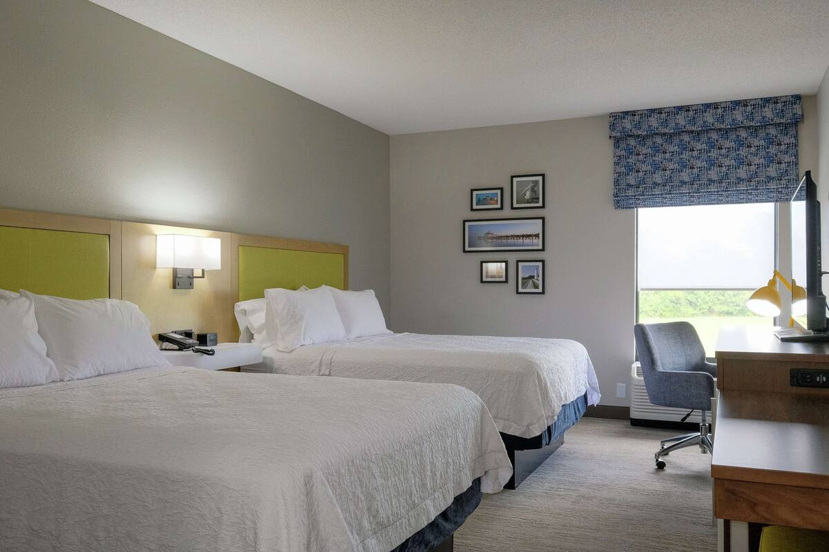 Habitación de hotel con dos camas, un escritorio con lámpara, una silla y una ventana con vistas.