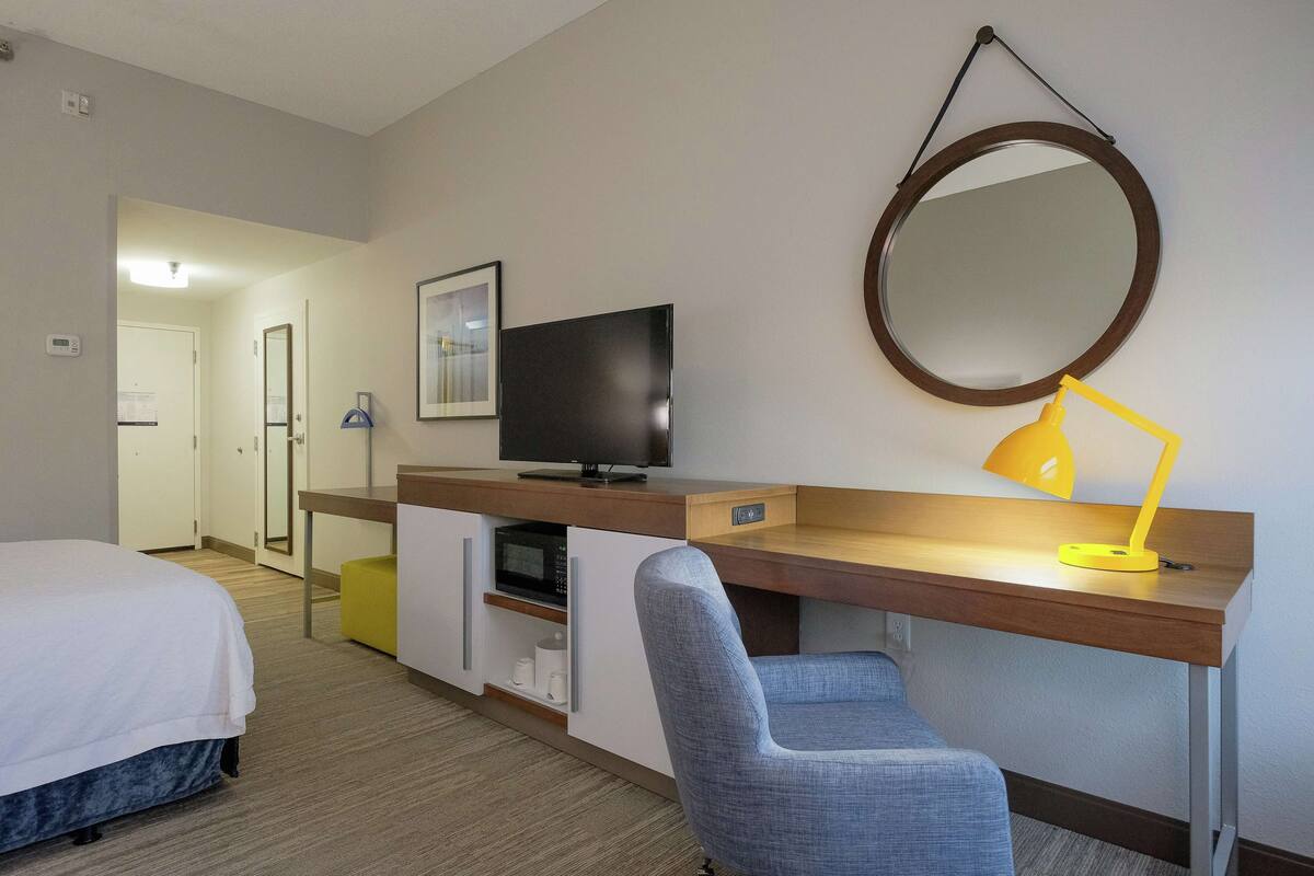 Habitación de hotel con cama, escritorio, computadora, silla, televisor, espejo y lámpara.