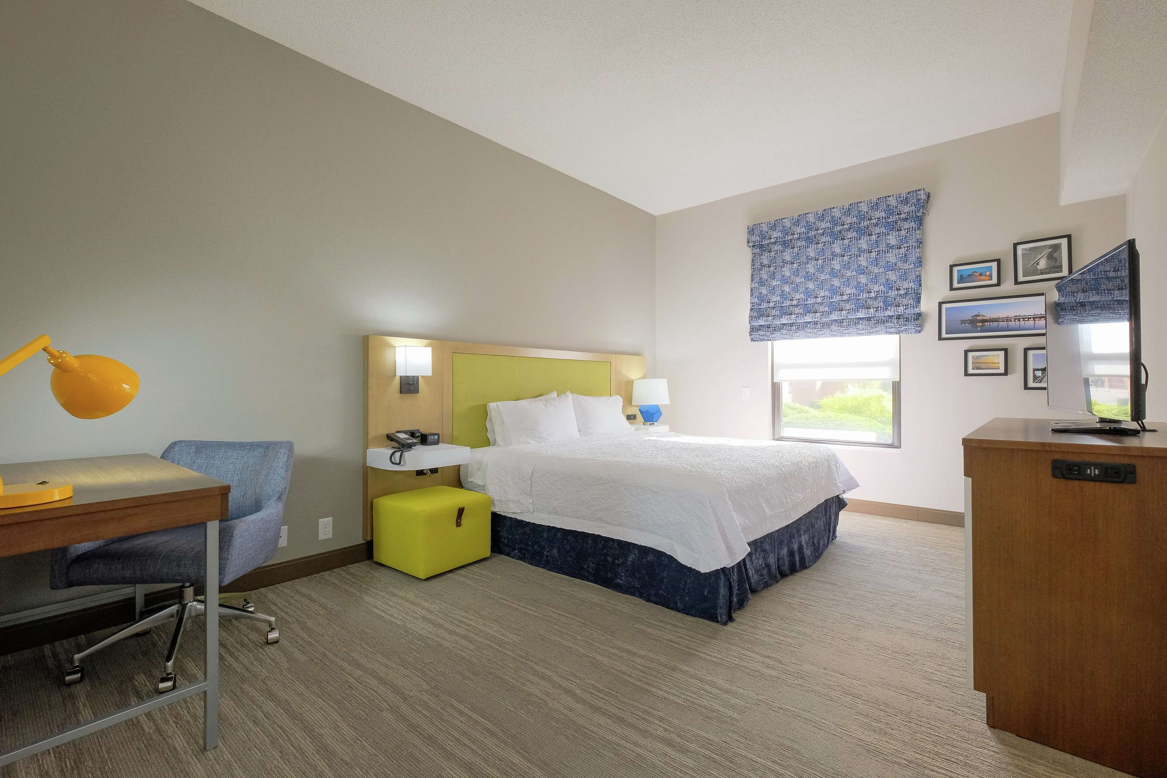 Habitación de hotel con cama, escritorio con silla, mesita pequeña, lámpara, mesita de noche, ventana con vistas y pared con cuadros.