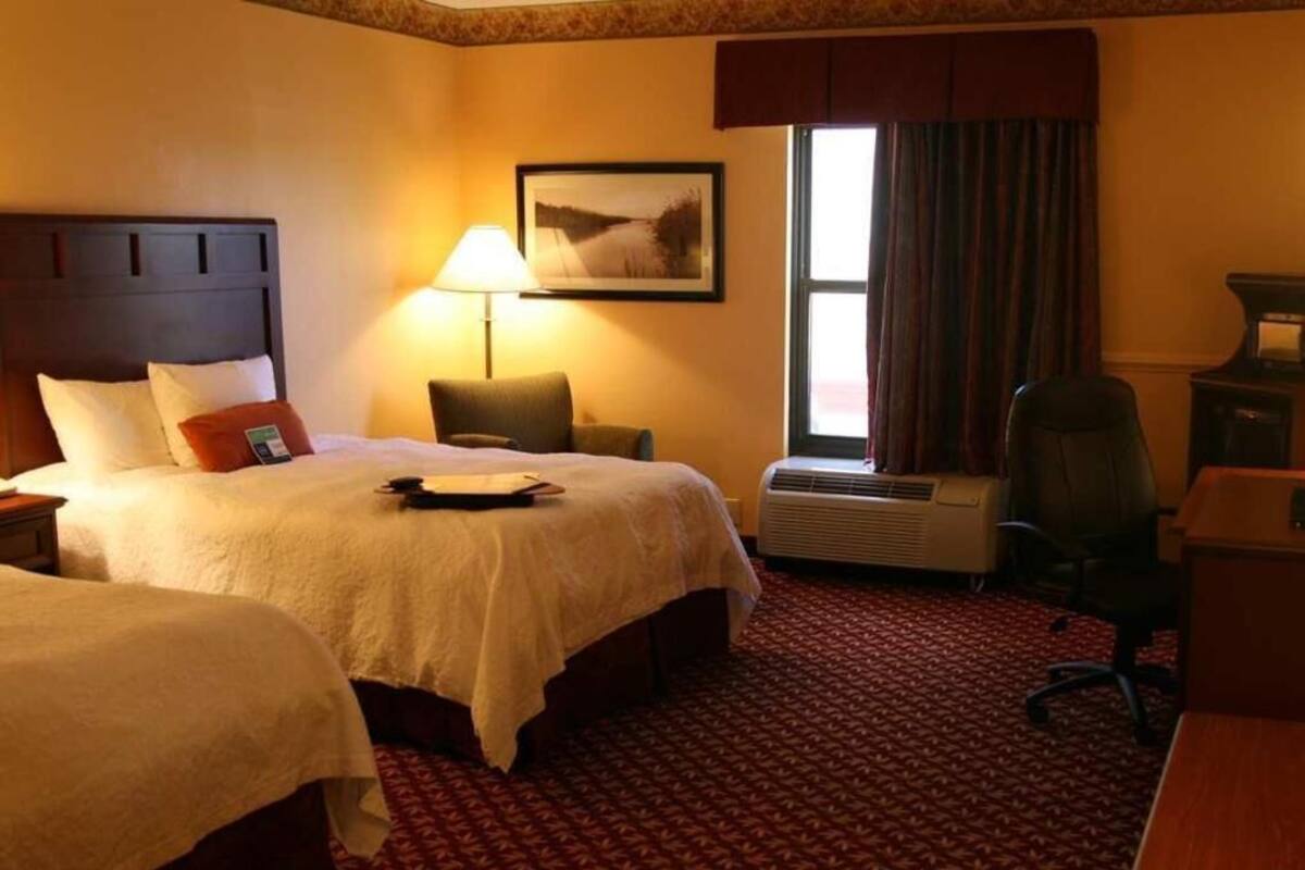 Habitación de hotel con dos camas, un escritorio, una silla, una lámpara, una ventana con cortinas y un equipo de aire acondicionado.