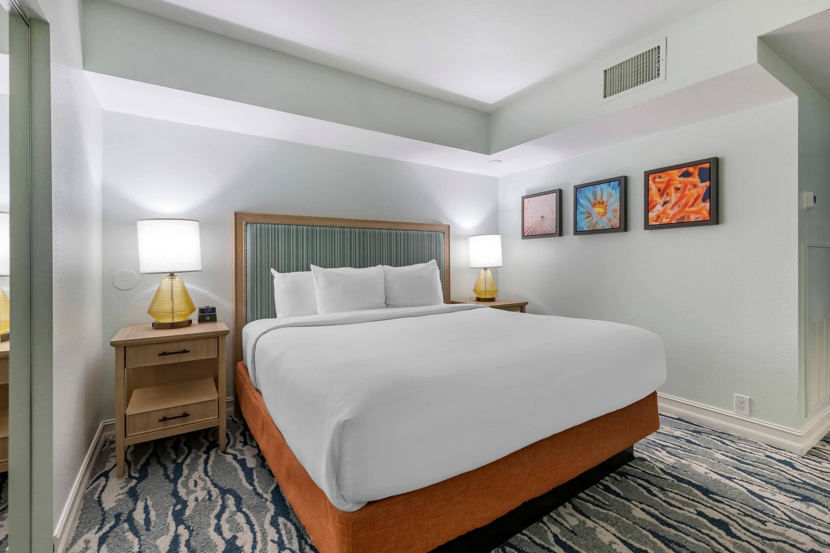 A hotel room with a large bed, two bedside tables with lamps, a small drawer unit, and three framed pictures on the wall.