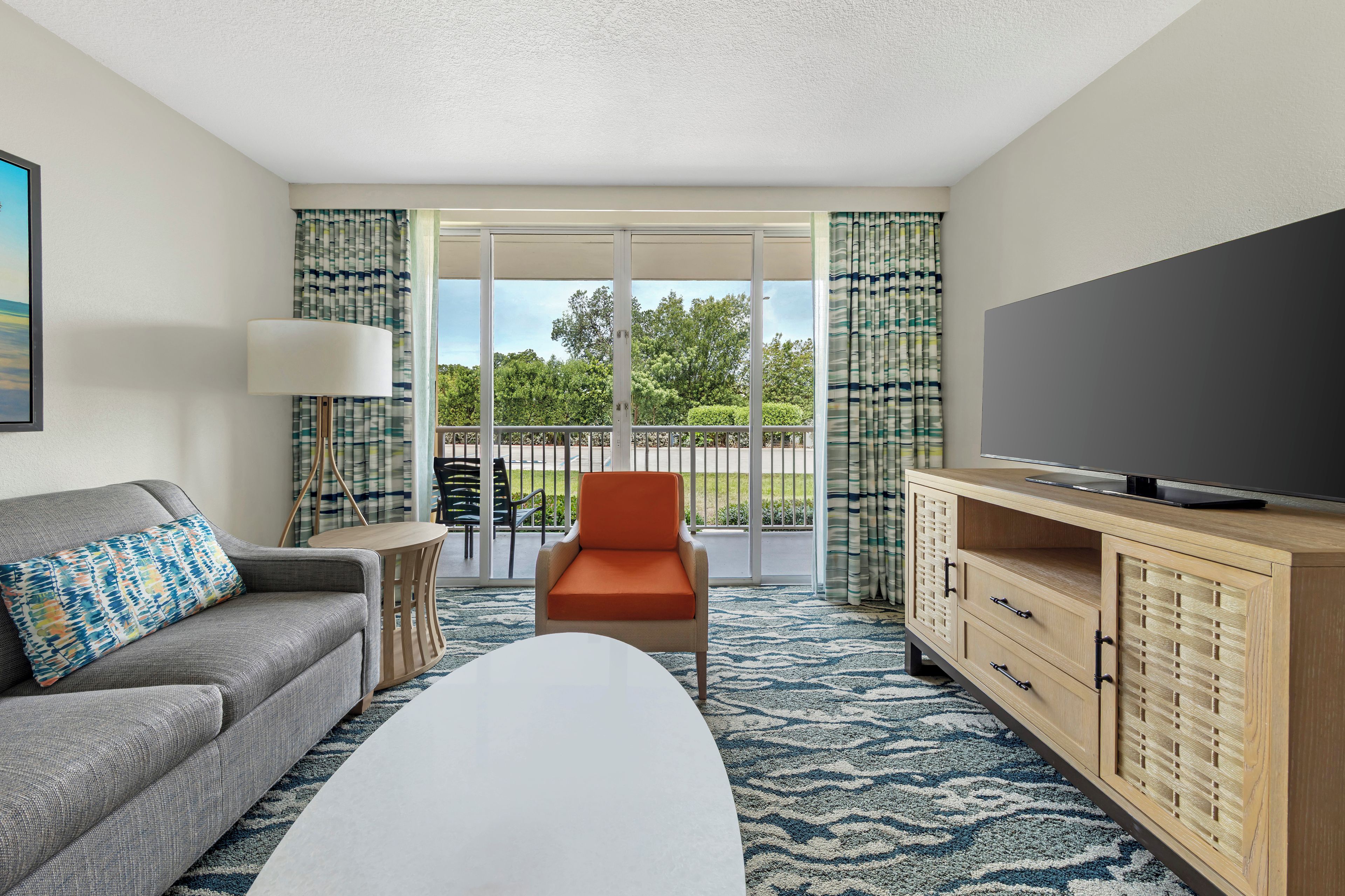 A hotel room with a sofa, a chair, a TV, a cabinet, and a balcony with a view of trees.