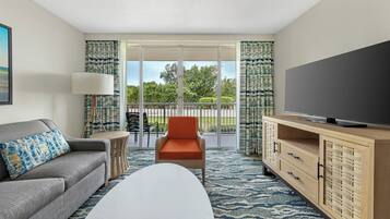 Habitación de hotel con sofá, sillón, televisor, armario y balcón con vistas a árboles.