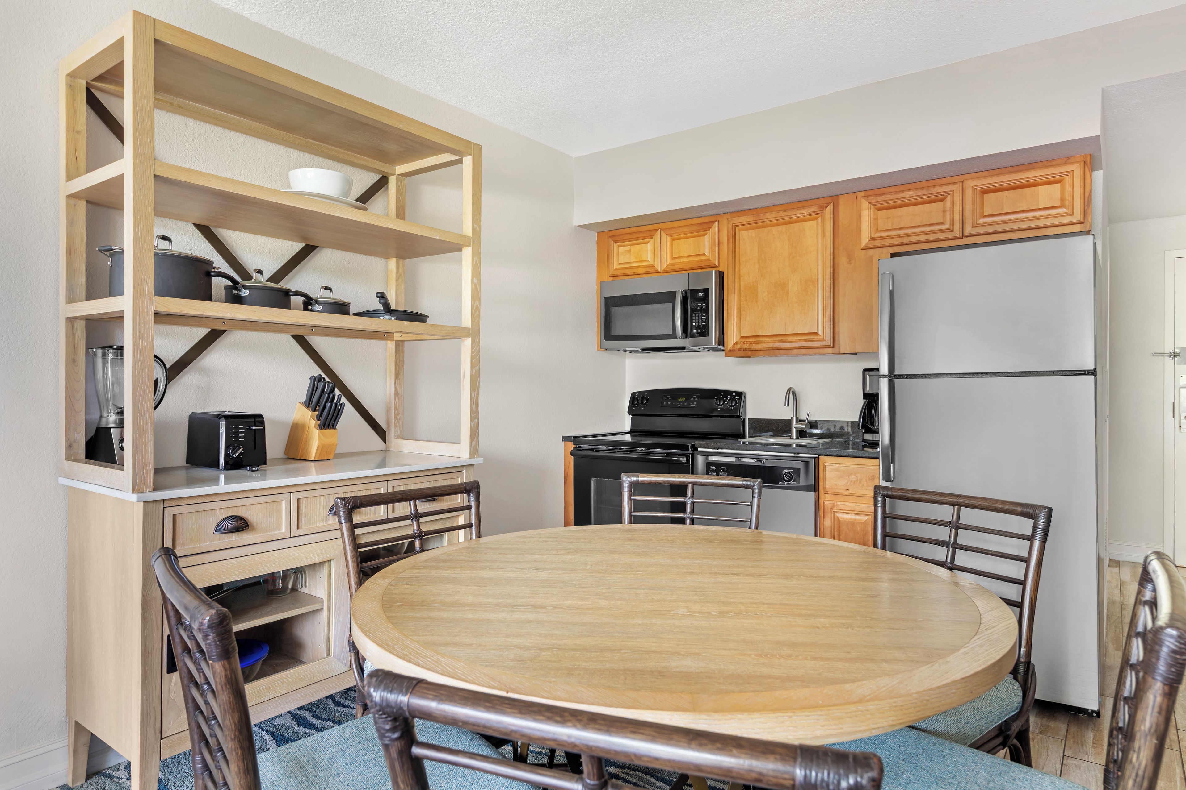 A compact kitchen with wooden cabinets, a microwave, a refrigerator, a dining table with chairs, and a built-in shelf unit with cookware.