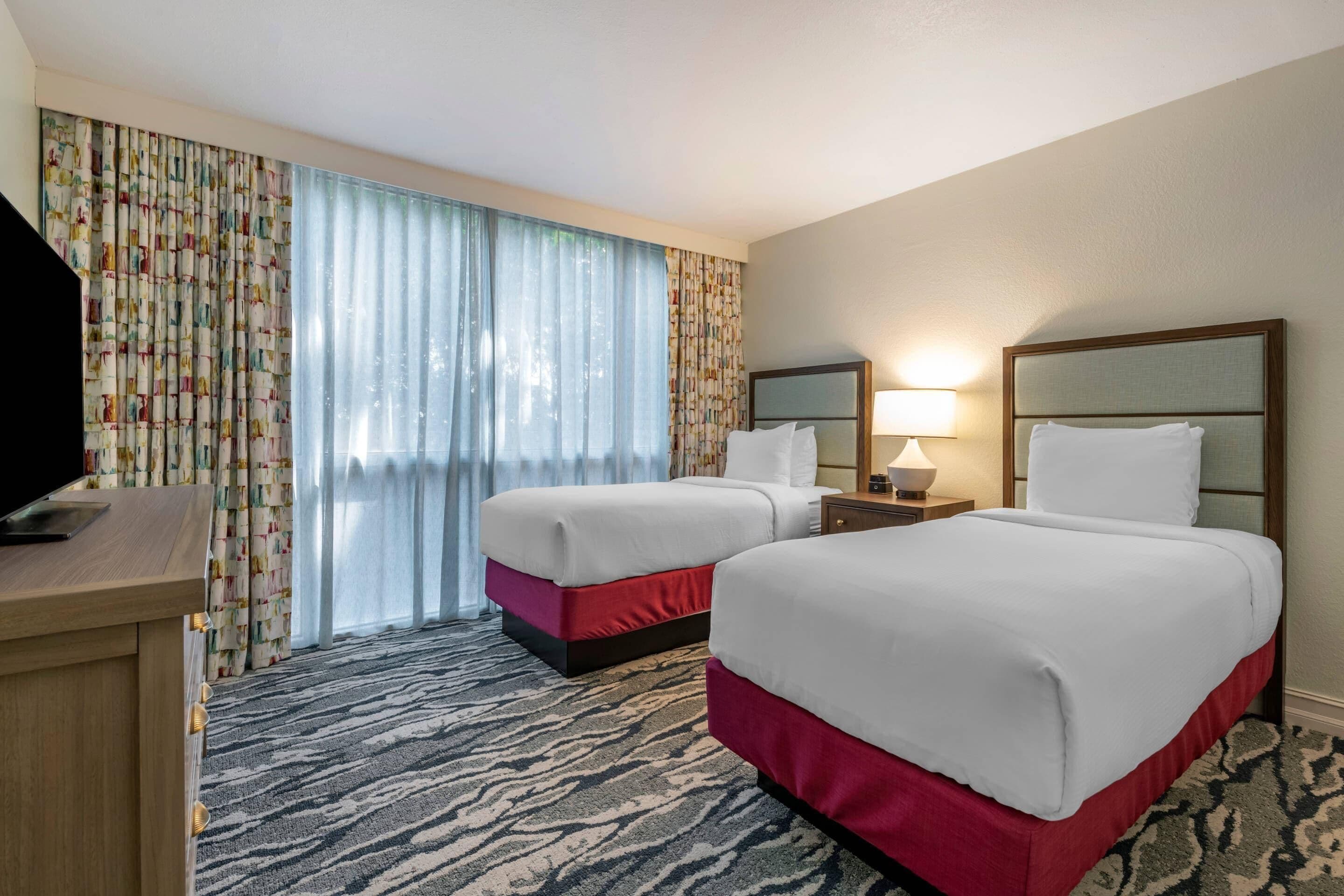 Quarto de hotel com duas camas, uma escrivaninha, uma luminária e uma televisão.