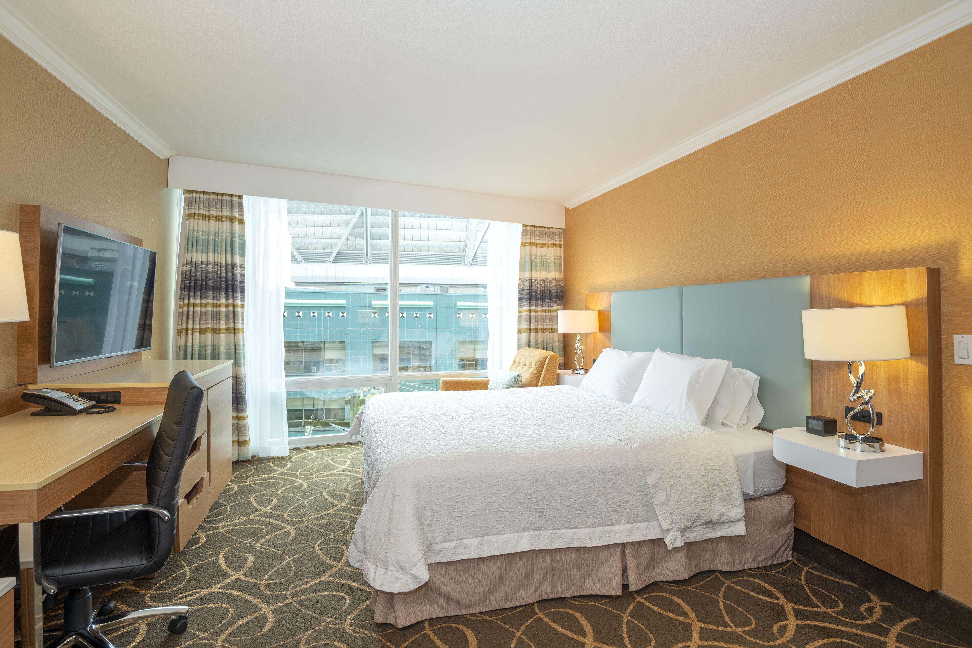 A hotel room with a bed, desk, chair, TV, and a view of a building through the window.