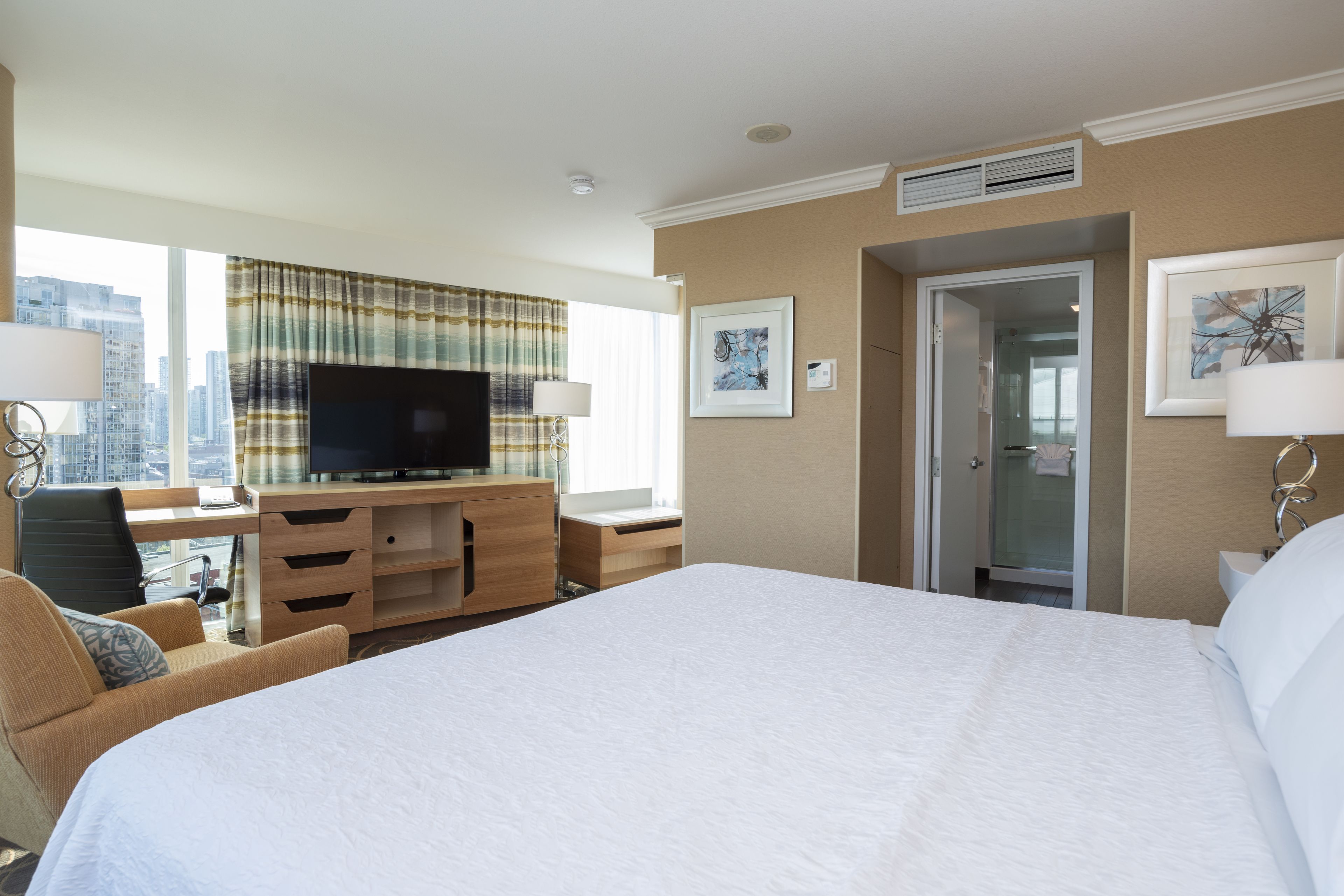 A hotel room with a large bed, a desk with a TV, a chair, and a view of the city.