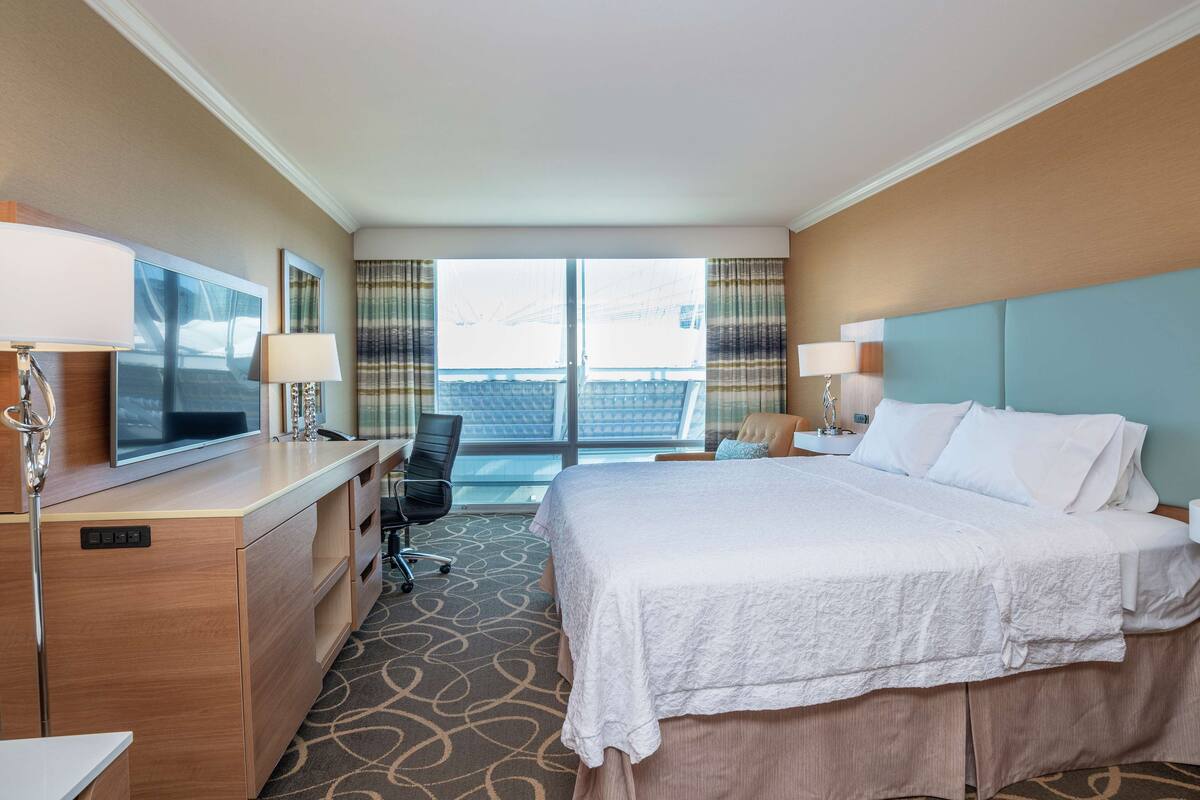 Habitación de hotel con cama, escritorio, televisión y vistas a la ciudad a través de amplios ventanales.