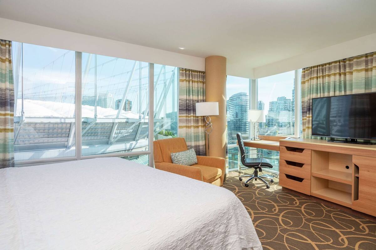 Habitación de hotel con una cama grande, un escritorio con televisor, una silla y vistas a edificios de la ciudad.