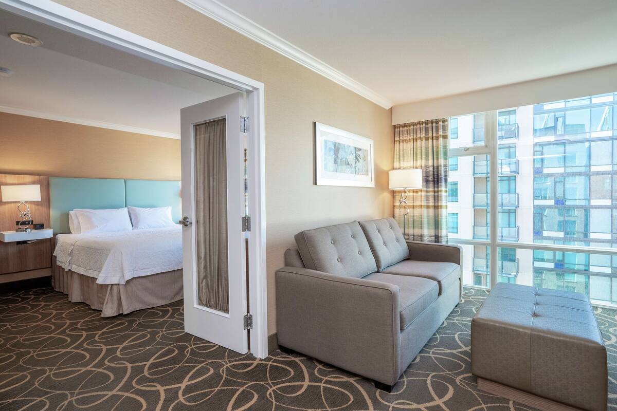 Habitación de hotel con cama, sofá, mesita y ventana con vista a edificios.
