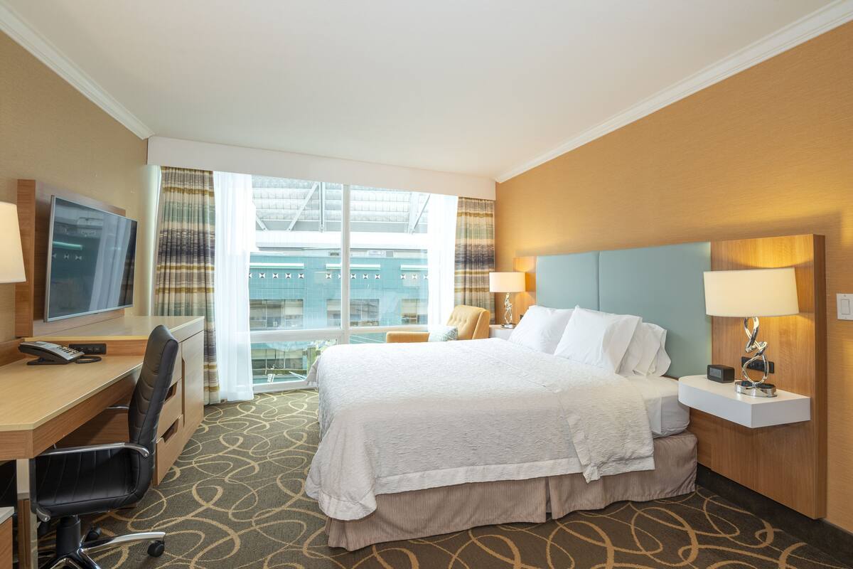 Habitación de hotel con cama, escritorio, silla, televisor y vista a un edificio a través del ventanal.