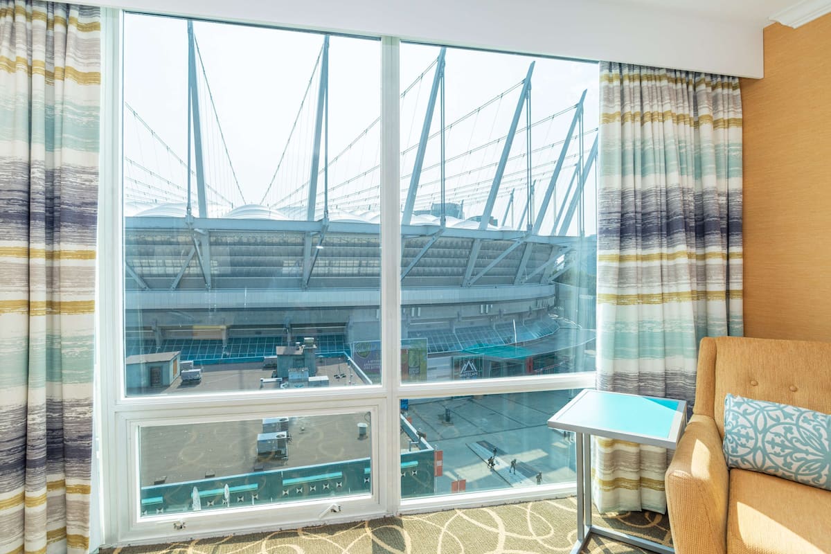 Habitación de hotel con un ventanal grande que ofrece vistas a un puente y un estadio. Hay una mesita pequeña y un sofá con un cojín estampado.