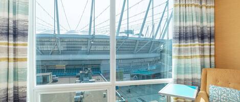 A hotel room with a large window offering a view of a bridge and a stadium. There is a small table and a sofa with a patterned pillow.