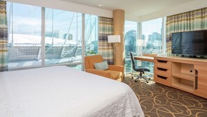 Ein Hotelzimmer mit einem großen Bett, einem Schreibtisch mit Computer, einem Stuhl, einem Fernseher und großen Fenstern mit Blick auf die Stadt.