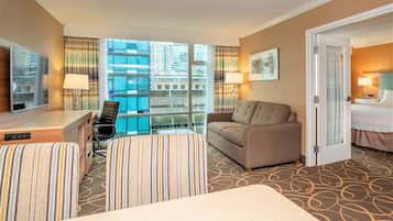 Quarto de hotel com cama, sofá, escrivaninha com cadeira, televisão e uma janela grande com vista para a cidade.