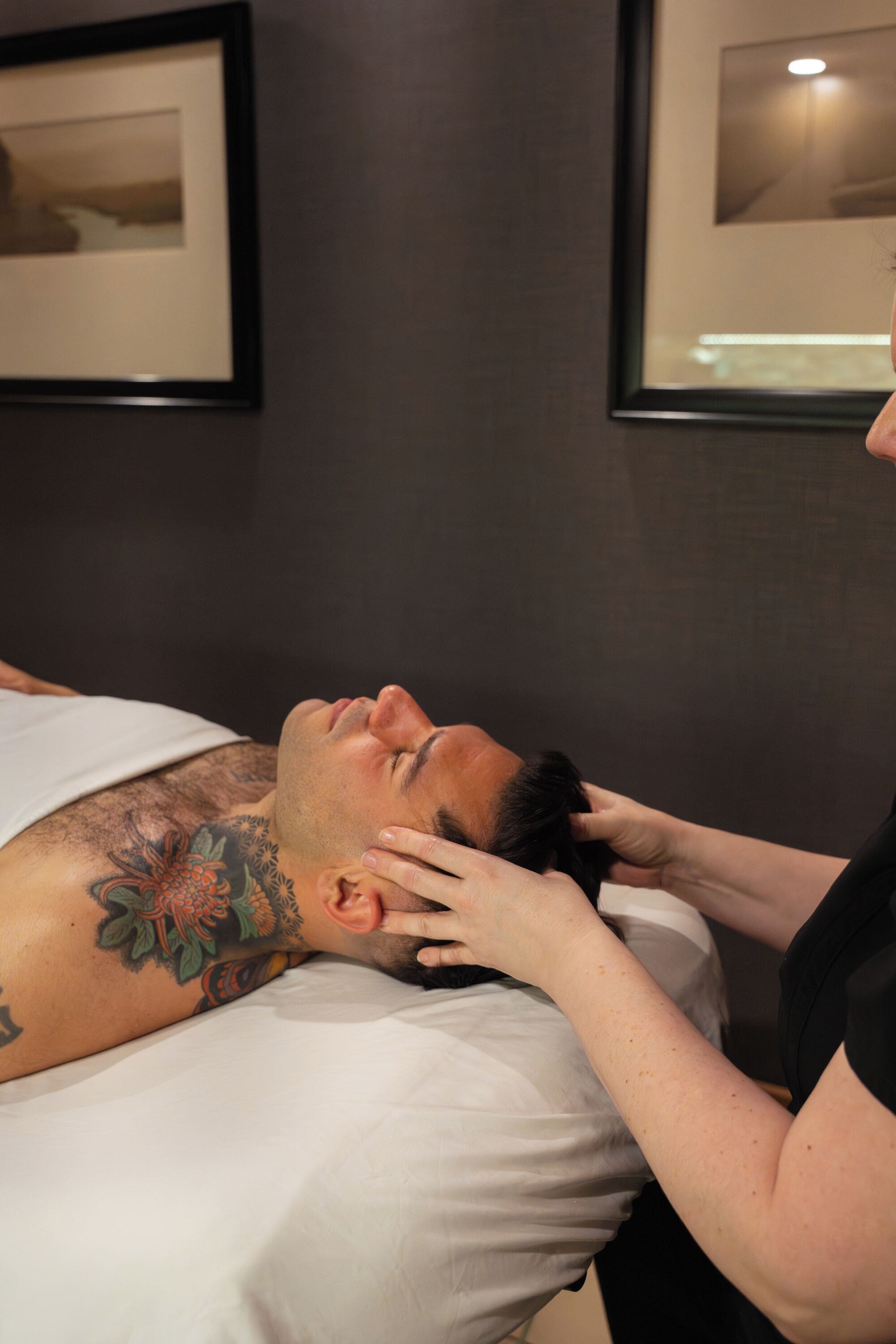 A person lying on a bed with a tattoo on their arm, in a room with framed pictures on the wall.