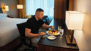A hotel room with a bed, a dining table with a plate of food, a glass of water, and a lamp.